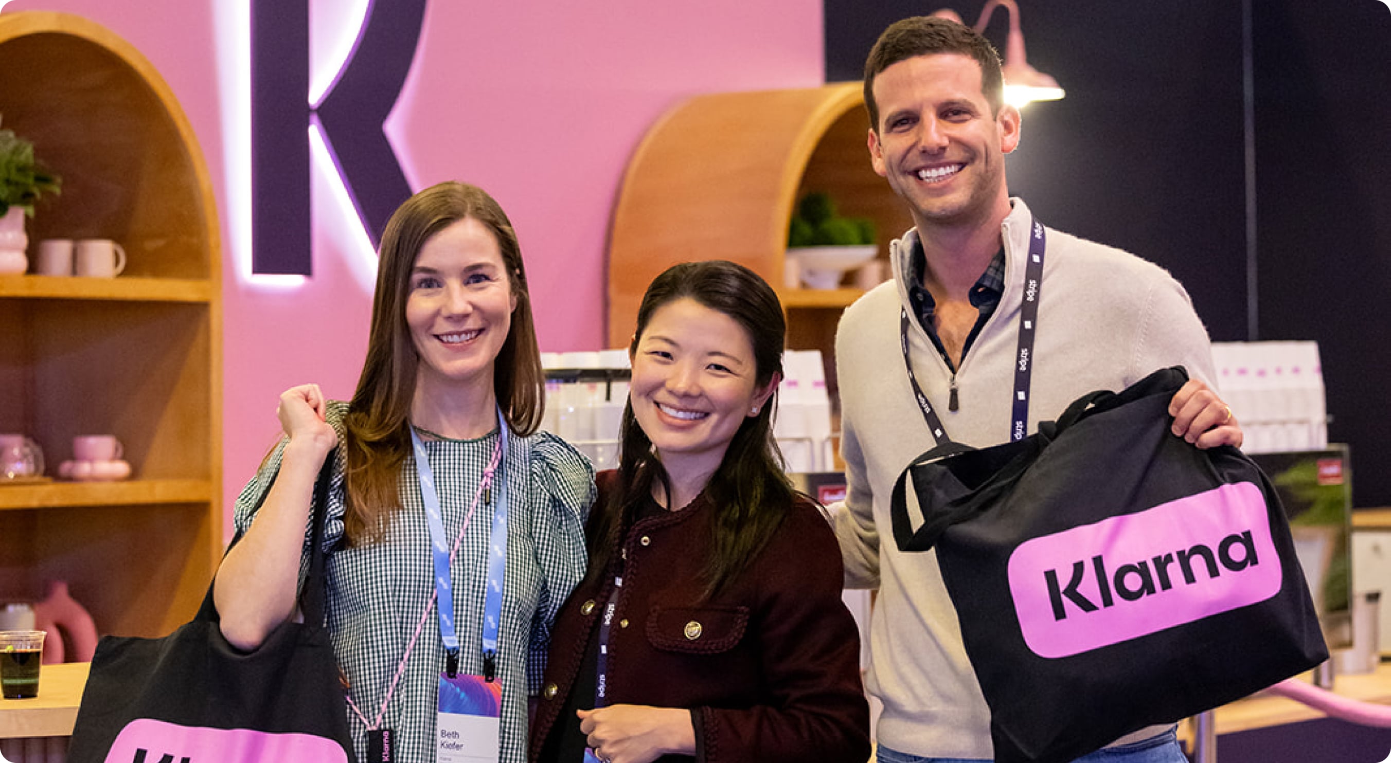 Group of sessions attendees holding Klarna canvas bags and smiling for the camera in front of the Klarna booth at sessions.